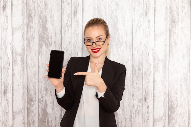 Junge Geschäftsfrau der Vorderansicht in der schwarzen Jacke der strengen Kleidung, die ihr Telefon auf weißer Oberfläche hält