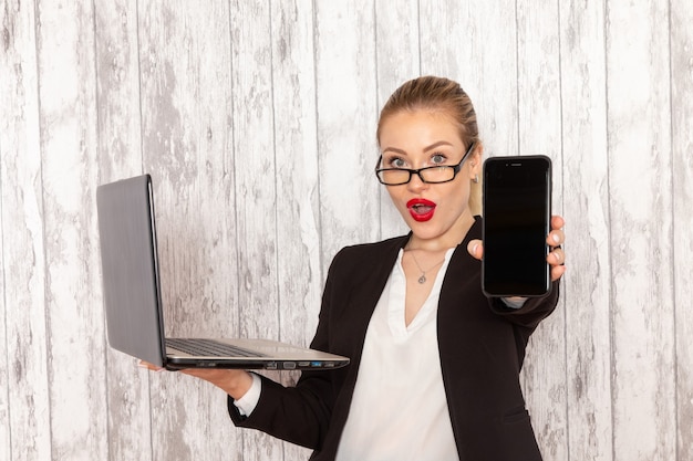 Kostenloses Foto junge geschäftsfrau der vorderansicht in der schwarzen jacke der strengen kleidung, die ihren laptop benutzt und telefon auf weißer oberfläche hält