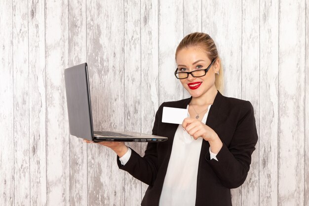 Junge Geschäftsfrau der Vorderansicht in der schwarzen Jacke der strengen Kleidung unter Verwendung ihres Laptops und der Haltekarte auf hellweißer Oberfläche