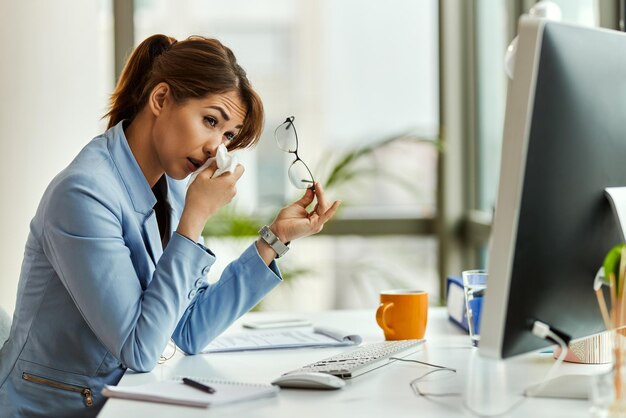 Junge Geschäftsfrau, die an einer Allergie leidet und Gesichtstücher verwendet, während sie an einem Computer im Büro arbeitet