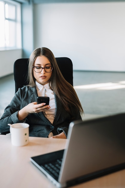 Junge Geschäftsfrau, die bei Tisch Smartphone mit Laptop verwendet