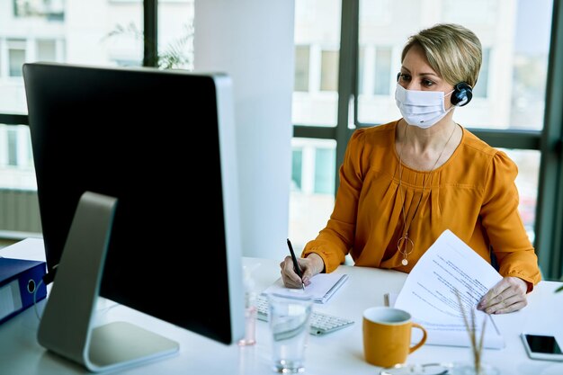Junge Geschäftsfrau, die eine schützende Gesichtsmaske trägt, während sie an einem Computer arbeitet und sich Notizen im Büro macht
