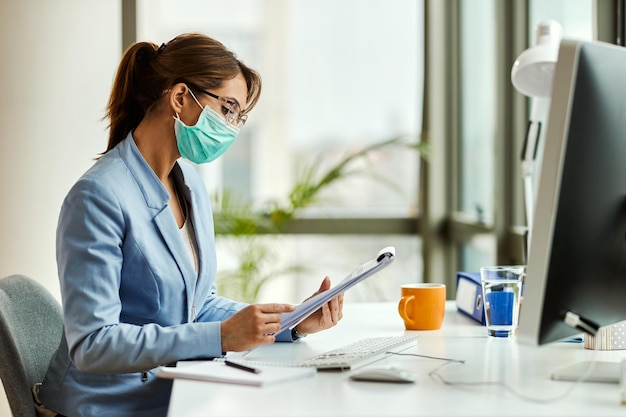 Junge Geschäftsfrau, die eine schützende Gesichtsmaske trägt, während sie Berichte durchgeht und im Büro arbeitet