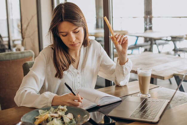 Junge Geschäftsfrau, die in einem Café zu Mittag isst und am Computer arbeitet