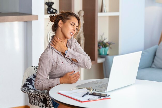 Junge Geschäftsfrau, die unter Nackenschmerzen leidet und ihren Nacken massiert, während sie an ihrem Arbeitsplatz im Home Office sitzt