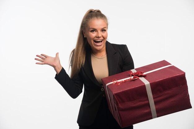 Junge Geschäftsfrau im schwarzen Anzug hält eine große Geschenkbox und sieht fröhlich aus.