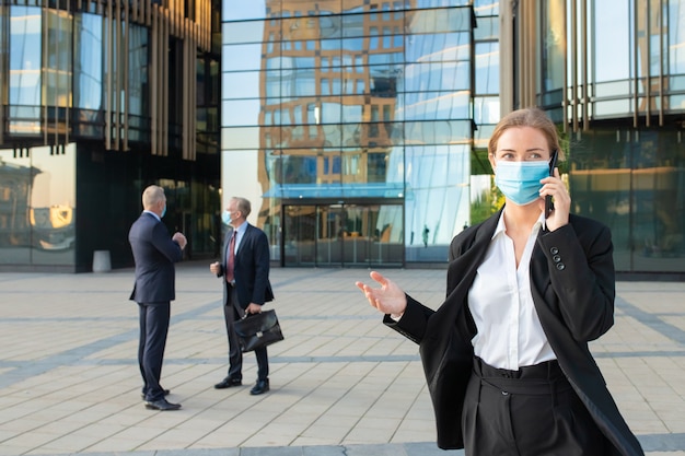 Kostenloses Foto junge geschäftsfrau in gesichtsmaske und büroanzug, die auf handy draußen spricht. geschäftsleute und stadtgebäude im hintergrund. speicherplatz kopieren. geschäfts- und epidemiekonzept