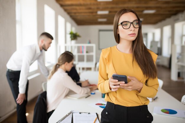 Junge Geschäftsfrau mit Brille schaut nachdenklich zur Seite, während sie im Büro mit Kollegen im Hintergrund das Handy in den Händen hält