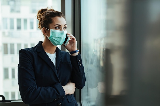 Junge Geschäftsfrau mit Gesichtsmaske, die am Fenster steht und telefoniert