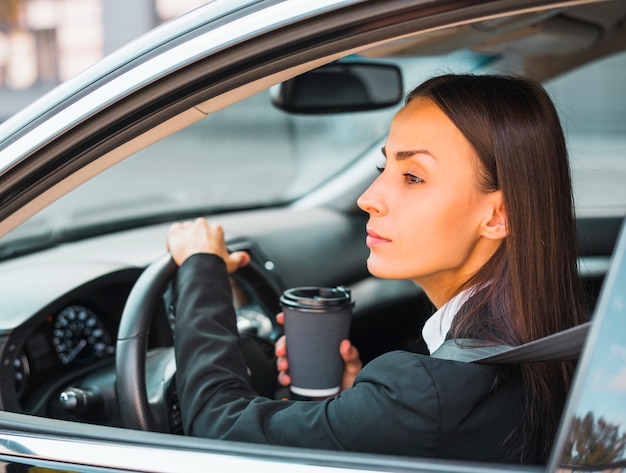 Junge Geschäftsfrau, welche die Wegwerfkaffeetasse fährt das Auto hält