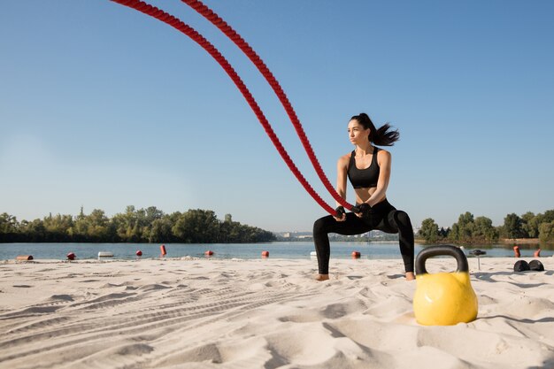 Junge gesunde Frau, die Übung mit den Seilen am Strand tut.