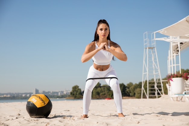 Junge gesunde Frau, die Kniebeugen mit dem Ball am Strand tut.