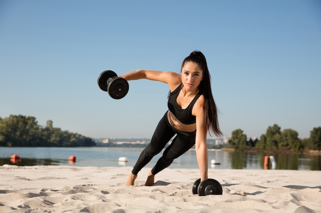 Junge gesunde Frau, die Oberkörper mit Gewichten am Strand trainiert