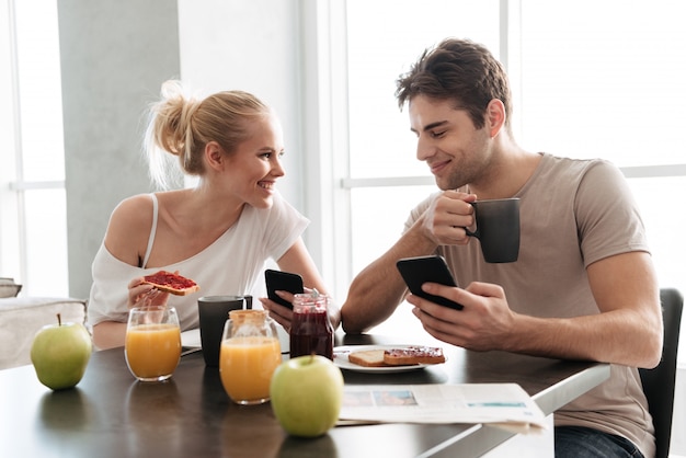 Junge gesunde Paare unter Verwendung ihrer Smartphones beim Essen des Frühstücks