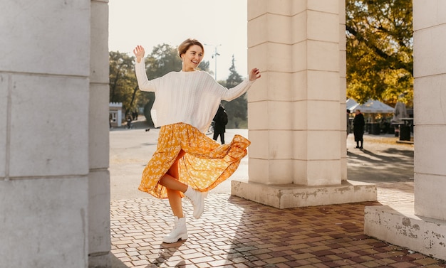 Junge glücklich lächelnde Frau in gelb bedrucktem Kleid und gestricktem weißen Pullover an einem sonnigen Herbsttag, der Spaß auf der Straße hat