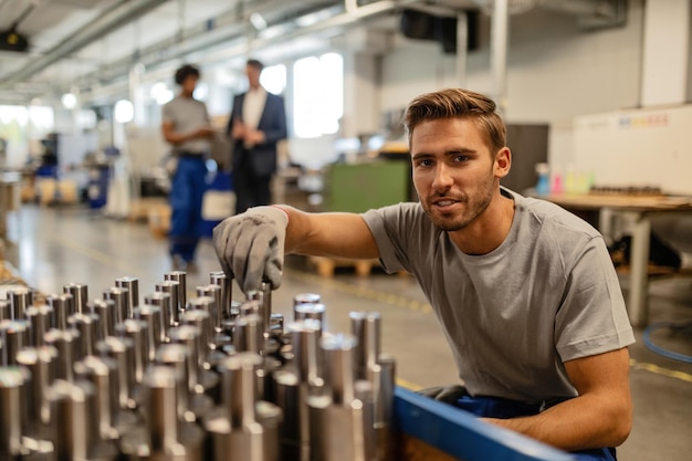 Junge glückliche Arbeiter, die die Qualität von Stahlstangen analysieren, während sie in Industriegebäuden arbeiten und in die Kamera schauen