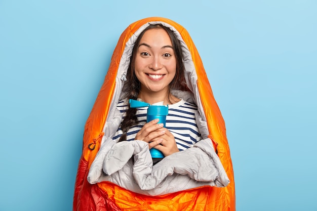 Junge glückliche asiatische Frau wärmt sich im Schlafsack, hält blaue Flasche mit heißem aromatischem Getränk, fühlt sich entspannt, verbringt Freizeit auf Natur, isoliert über blauer Wand