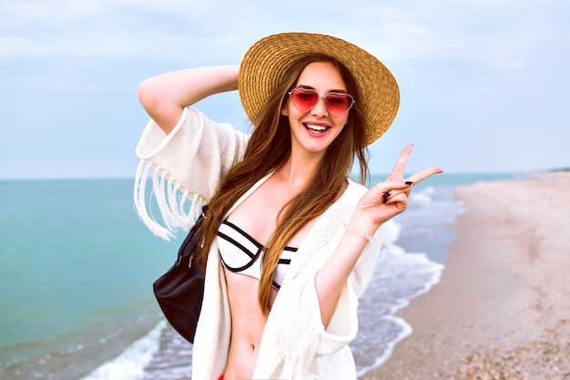 Junge glückliche blonde Mädchen posiert am Strand, trägt Strohhut und Herz niedliche Sonnenbrille, genießen Sie ihre Sommerferien in der Nähe des Ozeans, tragen Bikini und Boho-Jacke.