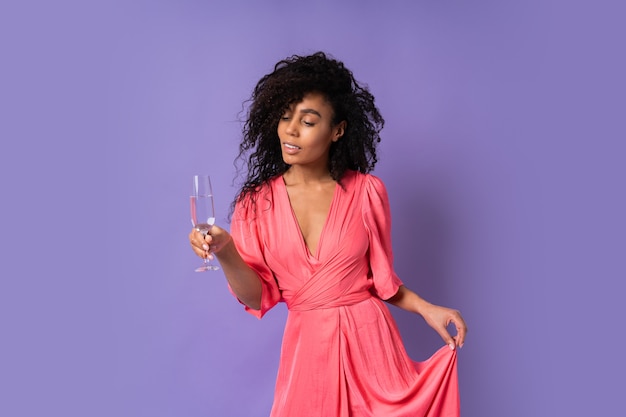 Junge glückliche brazillianische Frau mit lockigem Haar im rosa stilvollen Kleid, das mit Glas Champagner über lila Wand aufwirft. Partystimmung.
