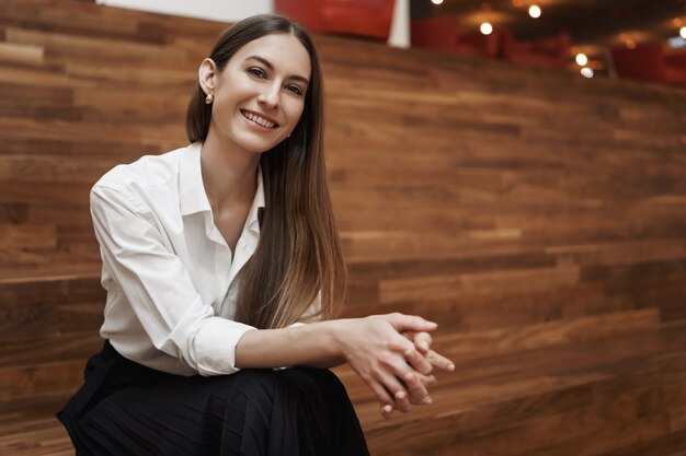 Junge glückliche erwachsene Frau in der Bluse und im langen Rock, die auf der Treppe innerhalb des Coworking Space sitzen und in die Kamera lächeln.