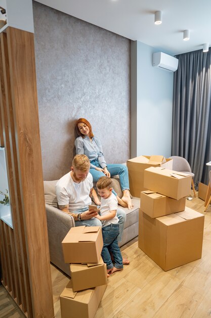 Junge glückliche Familie mit dem Kind, welches die Kästen zusammen sitzen auf Sofa auspackt