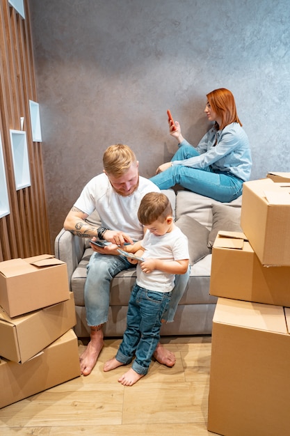 Junge glückliche familie mit dem kind, welches die kästen zusammen sitzen auf sofa auspackt