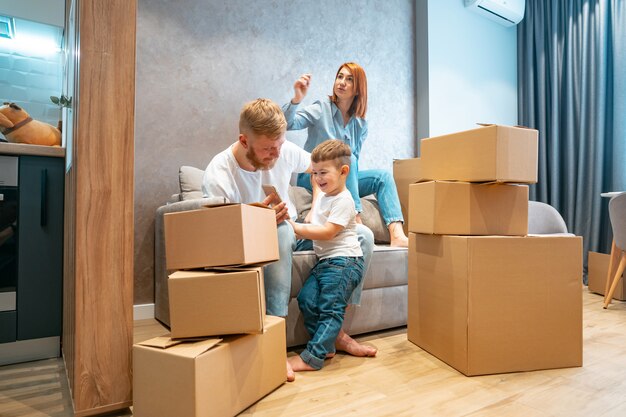 Junge glückliche Familie mit dem Kind, welches die Kästen zusammen sitzen auf Sofa auspackt