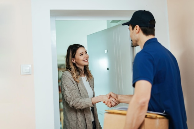 Junge glückliche Frau, die auf einer Tür steht und dem Lieferboten die Hand schüttelt, während sie ein Paket erhält