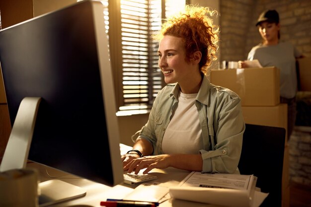 Junge glückliche Frau, die einen Computer benutzt, während sie eine E-Mail schreibt und im Büro des Kuriers arbeitet