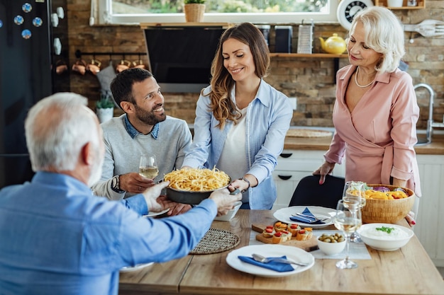 Junge glückliche Frau, die Essen am Esstisch serviert, während sie mit ihrem Mann und ihren reifen Eltern zu Hause zu Mittag isst