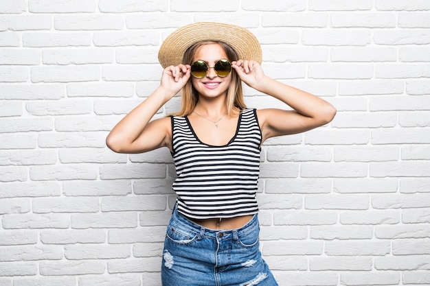 Kostenloses Foto junge glückliche frau, die in sonnenbrille und sommerhut über weiße backsteinmauer lächelt