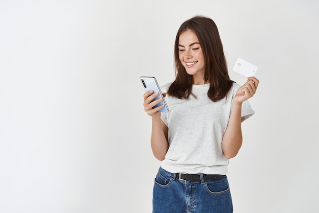 Junge glückliche frau, die mit smartphone und plastikkreditkarte zahlt, stehend über weißer wand.