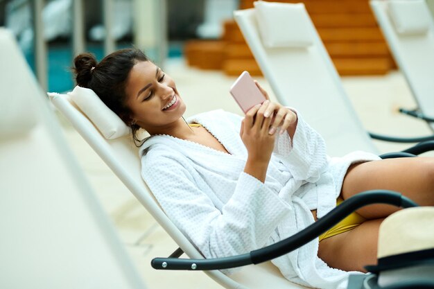 Junge glückliche Frau im Bademantel mit Smartphone beim Entspannen am Pool im Wellnesscenter