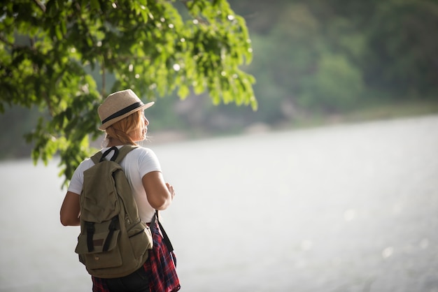 Junge glückliche Frau mit dem Rucksack, der schaut zum Fluss steht. Reisendes Konzept der Frau.