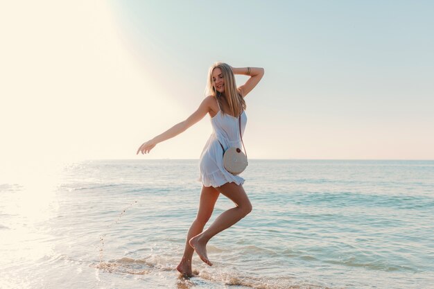 Junge glückliche Frau tanzt herum und dreht sich am sonnigen Sommermode-Stil des Seestrandes im weißen Kleid Urlaub