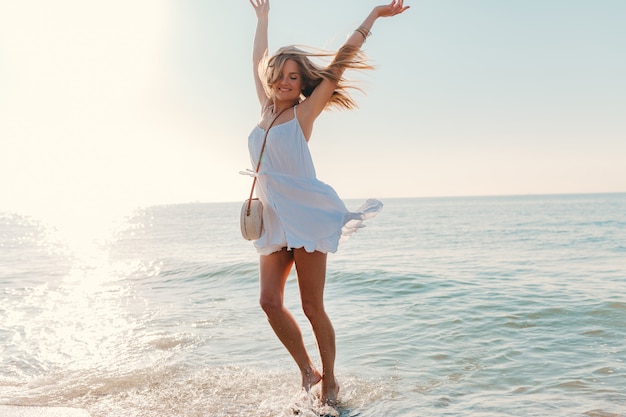 Junge glückliche Frau tanzt herum und dreht sich am sonnigen Sommermode-Stil des Seestrandes im weißen Kleid Urlaub
