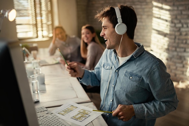 Junge glückliche Freiberufler, die Musik über Kopfhörer hören und Spaß haben, während sie im Büro arbeiten Seine Kollegen sind im Hintergrund