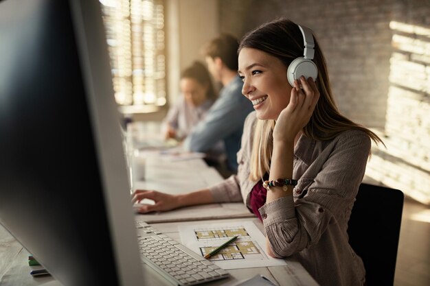 Junge glückliche Geschäftsfrau, die an einem Computer arbeitet und im Büro Musik über Kopfhörer hört. Ihre Kollegen arbeiten im Hintergrund