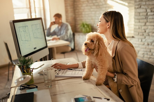 Junge glückliche Geschäftsfrau, die an einem Computer arbeitet, während sie mit ihrem Hund im Büro ist