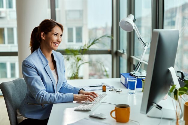 Junge glückliche Geschäftsfrau, die Desktop-PC verwendet, während sie im Büro arbeitet