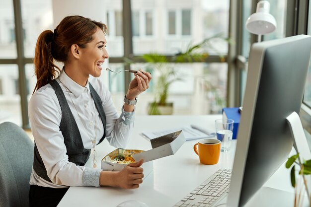 Junge glückliche Geschäftsfrau, die in einer Pause im Büro isst