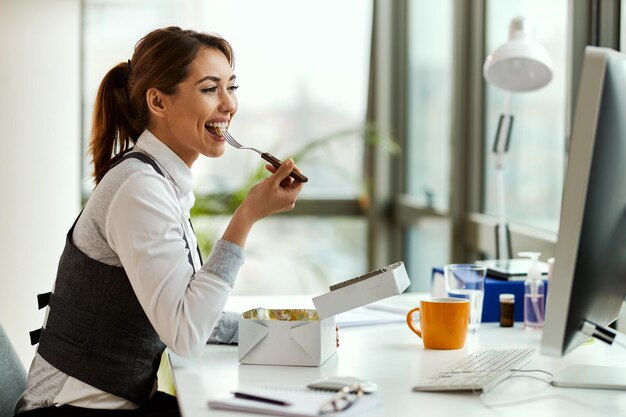 Junge glückliche Geschäftsfrau, die isst, während sie einen Desktop-PC im Büro verwendet