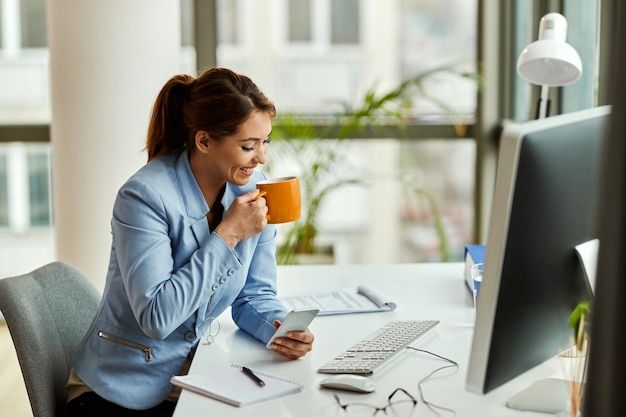 Junge glückliche Geschäftsfrau trinkt Kaffee und SMS auf dem Handy im Büro