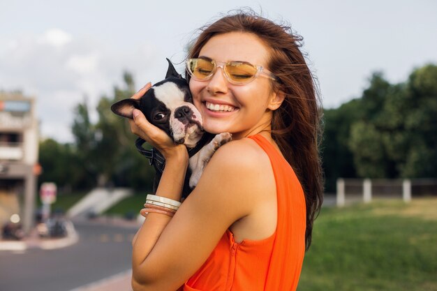 Junge glückliche lächelnde Frau, die Boston Terrrier Hund im Park, sonnigen Sommertag, fröhliche Stimmung hält, mit Haustier spielt, langes Haar winkt, Spaß hat, Sommermodetrend