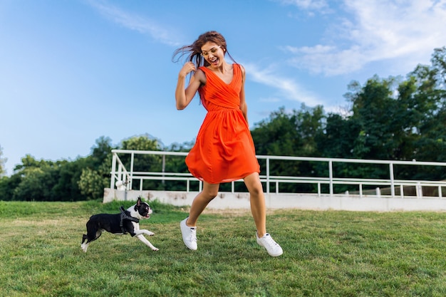 Junge glückliche lächelnde Frau im orangefarbenen Kleid, das Spaß beim Spielen mit Hund im Park, Sommerart, fröhliche Stimmung spielt