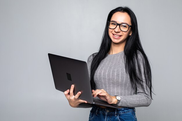 Junge glückliche lächelnde Frau in der Freizeitkleidung, die Laptop lokalisiert auf grauer Wand hält