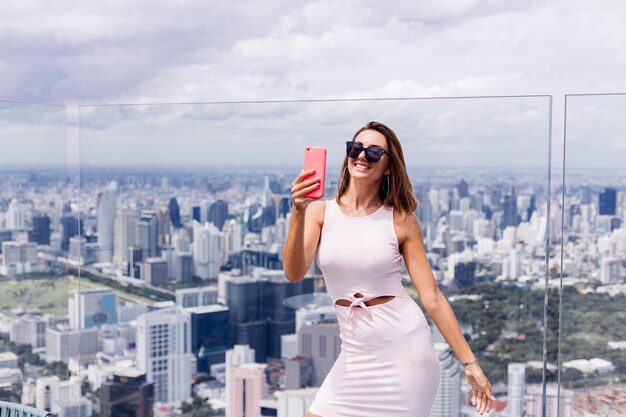 Junge glückliche lächelnde kaukasische Frau Reisende in passendem Kleid und Sonnenbrille auf hohem Boden in Bangkok halten Telefon