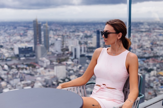 Junge glückliche lächelnde kaukasische Frau Reisende in passendem Kleid und Sonnenbrille auf hohem Boden in Bangkok Stilvolle Frau, die erstaunliche Großstadtansicht erkundet