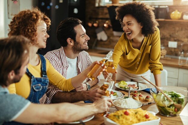 Junge glückliche Menschen, die Spaß haben und mit Bier anstoßen, während sie zu Hause im Speisesaal zu Mittag essen.