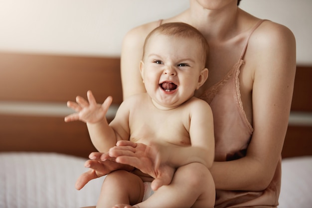 Junge glückliche Mutter in Nachtwäsche und ihr neugeborenes Baby sitzen am Morgen lächelnd auf dem Bett und spielen zusammen.